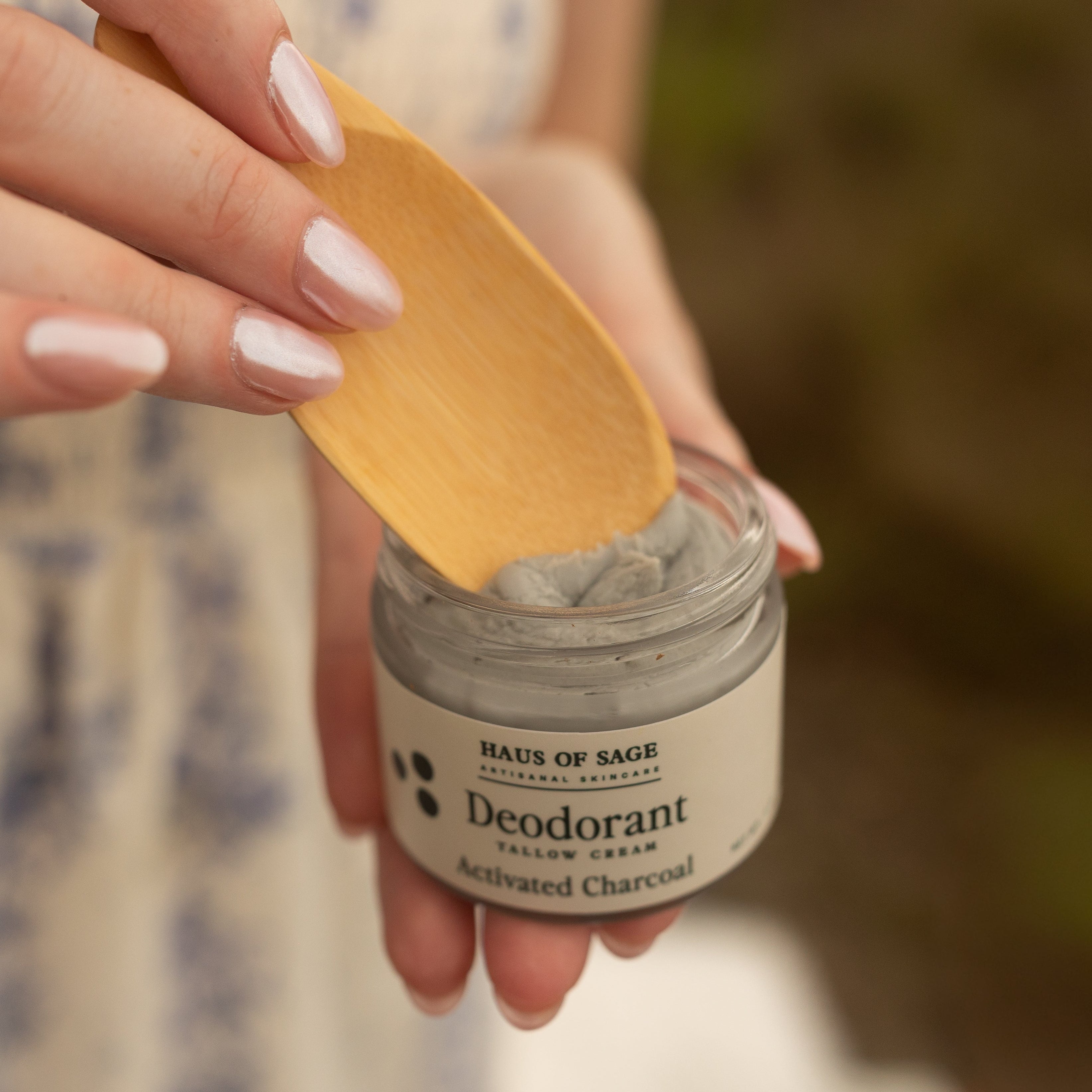 A close-up of a person holding a small jar of charcoal deodorant cream with a wooden spoon, wearing a cream-colored dress with a blue floral pattern against a blurred outdoor background.