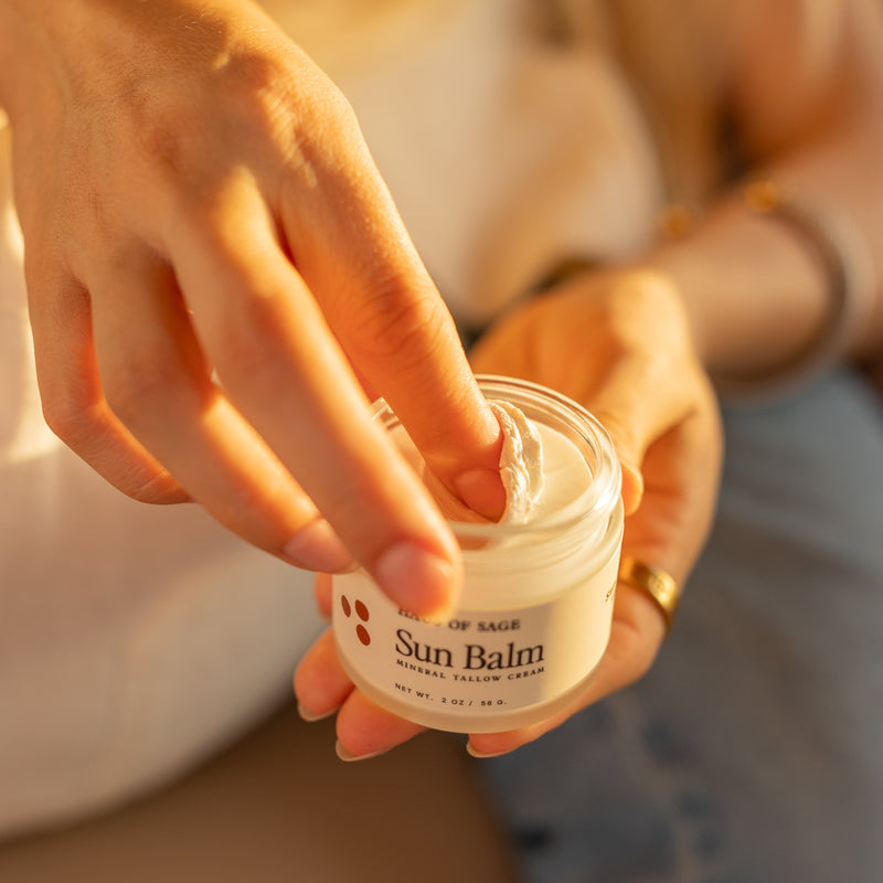 A close-up lifestyle shot of a person holding a small jar of sun balm, with their fingers touching the cream inside. The person is wearing a white top and blue jeans, and the background is softly blurred in warm sunlight.