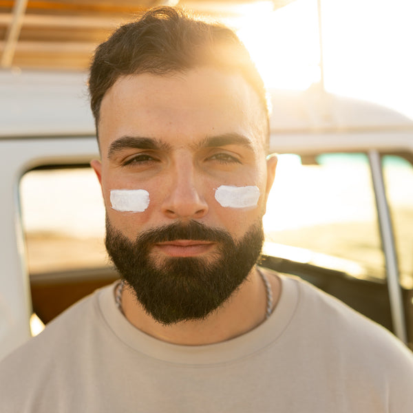 A close-up portrait of a man with a dark beard and short hair, wearing a light beige t-shirt and having white sunscreen blocks applied to his cheeks. He is positioned in front of a vintage-style white vehicle with warm, backlit sunlight creating a lens flare.