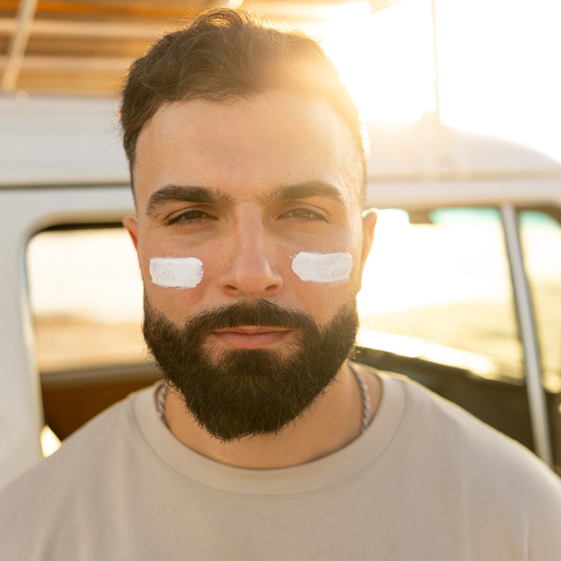 A close-up portrait of a man with a dark beard and short hair, wearing a light beige t-shirt and having white sunscreen blocks applied to his cheeks. He is positioned in front of a vintage-style white vehicle with warm, backlit sunlight creating a lens flare.