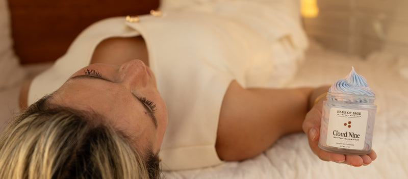 A woman lying on a bed holds a small jar of Blue Loo Soak feet bath salts. The jar has a white label with the brand name 'BLUE LOO SOAK' and the scent 'Cloud Nine', topped with a swirl of light blue bath salts.
