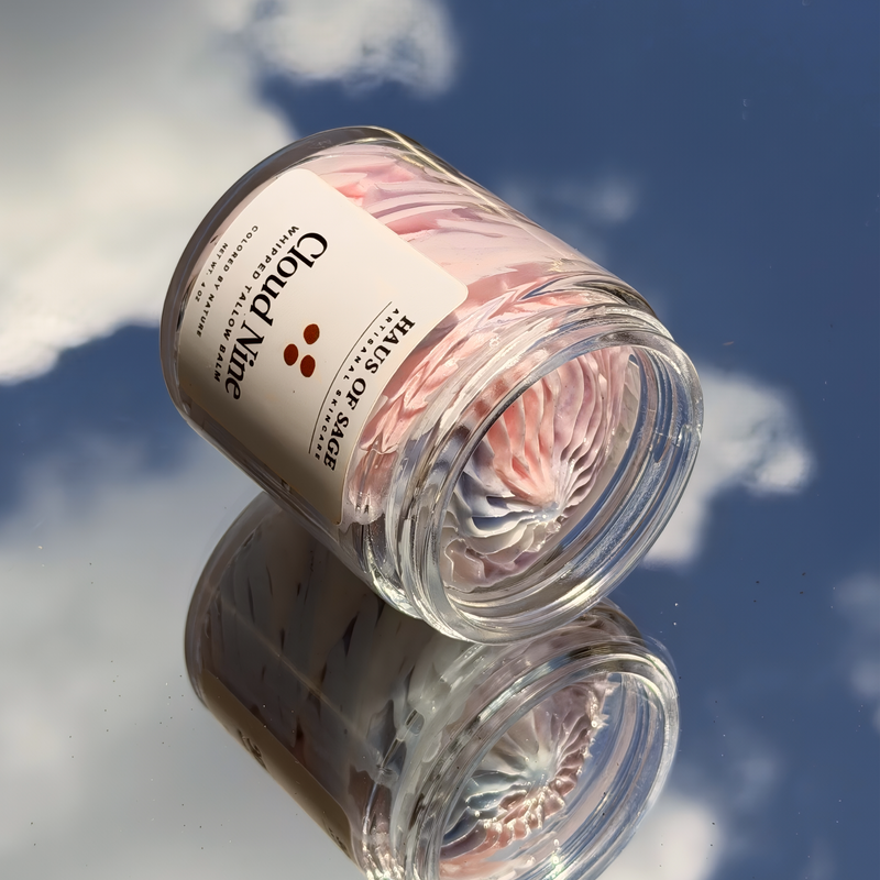 A clear glass jar candle with pale pink wax and a white label reading 'Cloud Mine' photographed on a reflective surface against a blue sky with clouds.