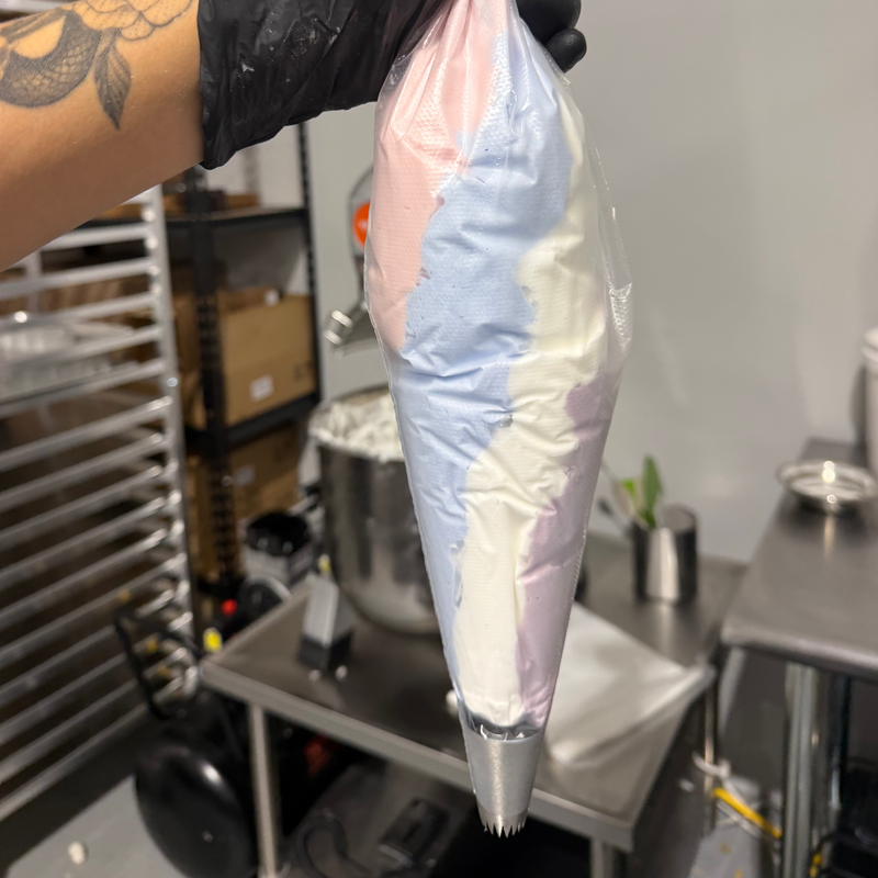 A hand wearing a black glove holds a clear plastic piping bag filled with pastel pink, blue, white, and lavender cream in a commercial kitchen setting.