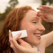 Woman applying sunscreen to her face with a hand holding a sunscreen bottle.